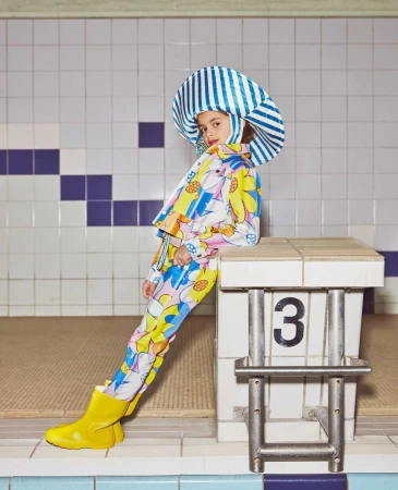 Child model poses next to a starting block, wearing a striped hat, floral jacket, pants, and yellow rain boots in an indoor pool area.