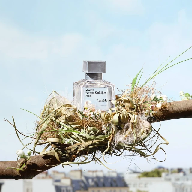Maison Francis Kurkdjian Petit Matin perfume bottle on a branch with botanical elements against a blurred cityscape and blue sky.