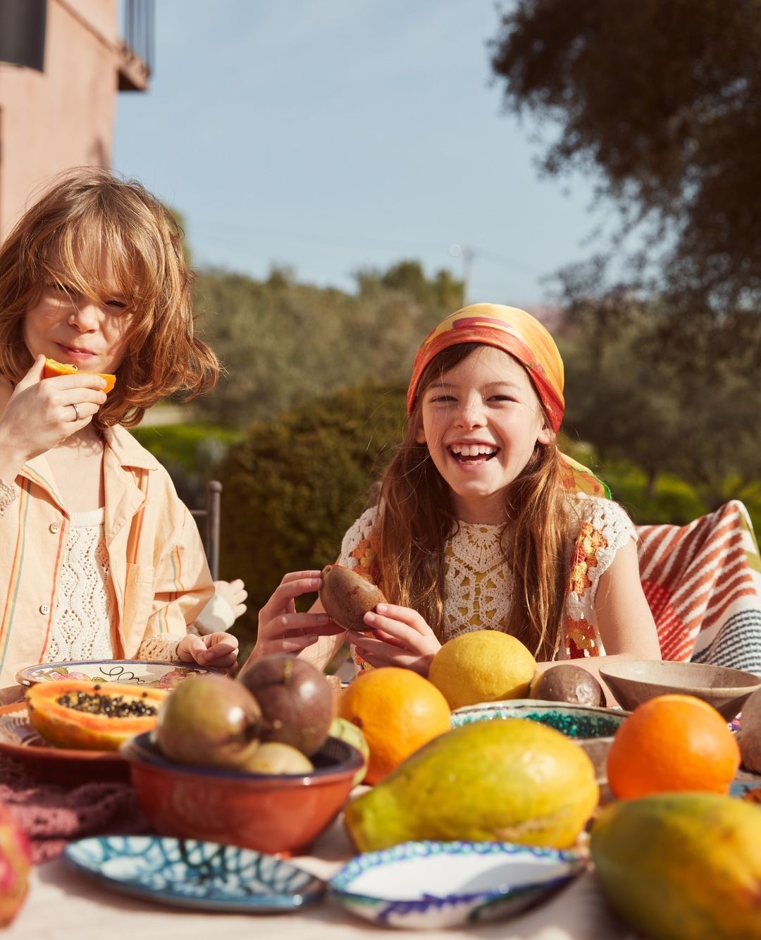 Two young girls enjoy a sunny outdoor meal with fresh fruit: oranges, passion fruit, pears, papaya, and pomegranates on a table.