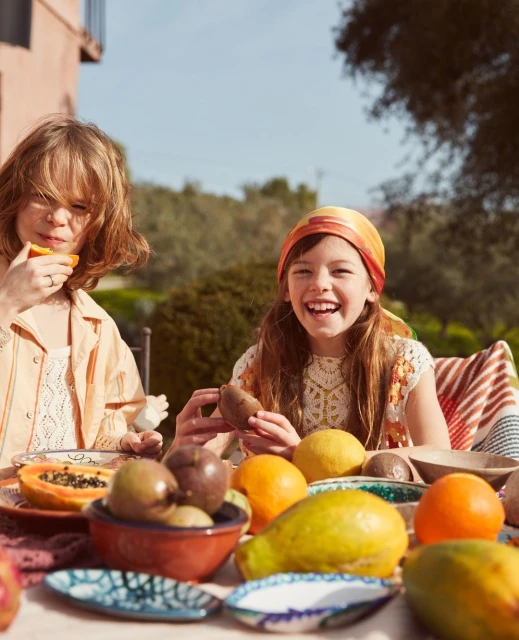 Two young girls enjoy a sunny outdoor meal with fresh fruit: oranges, passion fruit, pears, papaya, and pomegranates on a table.