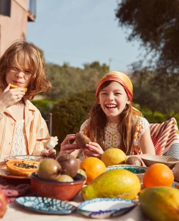 Two young girls enjoy a sunny outdoor meal with fresh fruit: oranges, passion fruit, pears, papaya, and pomegranates on a table.