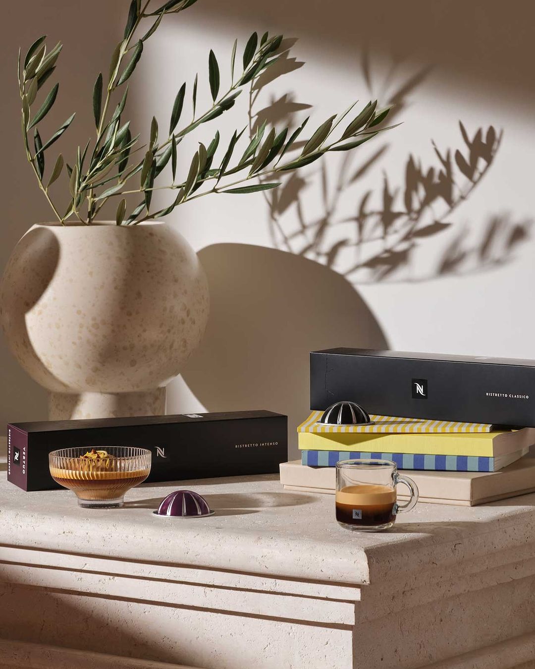 Still life featuring Nespresso coffee capsules, a coffee cup, and a vase with leafy branches, all arranged on a textured surface.