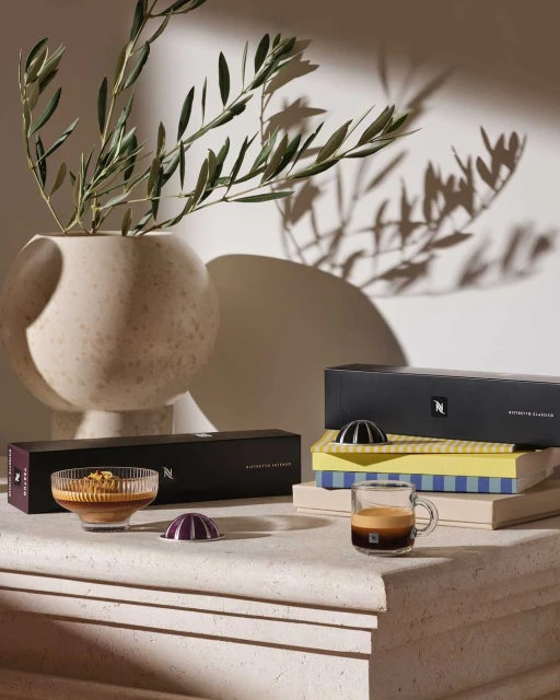 Still life featuring Nespresso coffee capsules, a coffee cup, and a vase with leafy branches, all arranged on a textured surface.