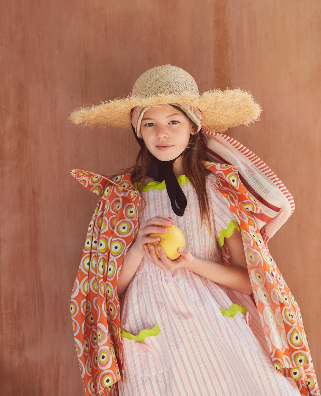 A young girl wearing a straw hat and a patterned dress holds a yellow fruit against a terracotta background, showcasing a whimsical and colorful aesthetic.