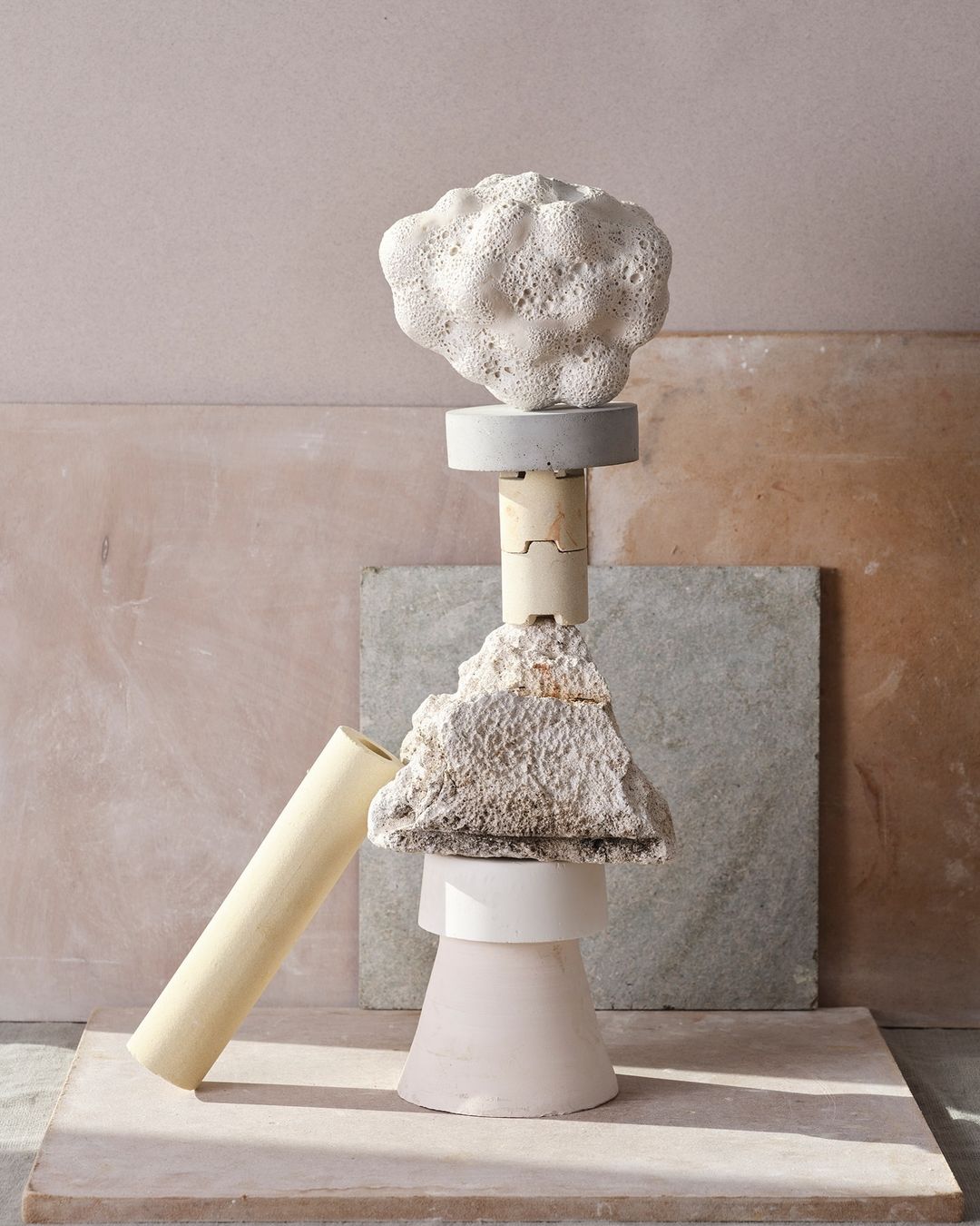 Abstract still life featuring sculptural arrangement of geometric shapes, rough stones, and cylinder against a textured backdrop.
