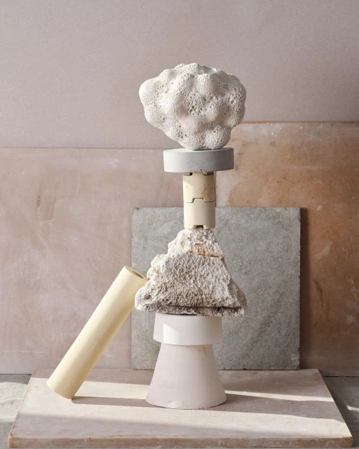 Abstract still life featuring sculptural arrangement of geometric shapes, rough stones, and cylinder against a textured backdrop.