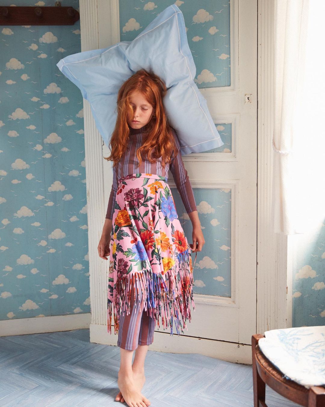 A young girl with red hair poses with a large pillow in an indoor setting with cloud-patterned wallpaper and a floral patterned skirt.