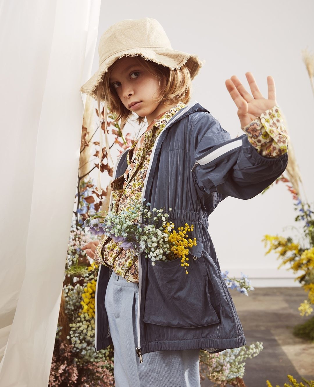 Young child in a blue jacket and floral shirt posing in front of a colorful flower background, wearing a bucket hat and holding up four fingers.