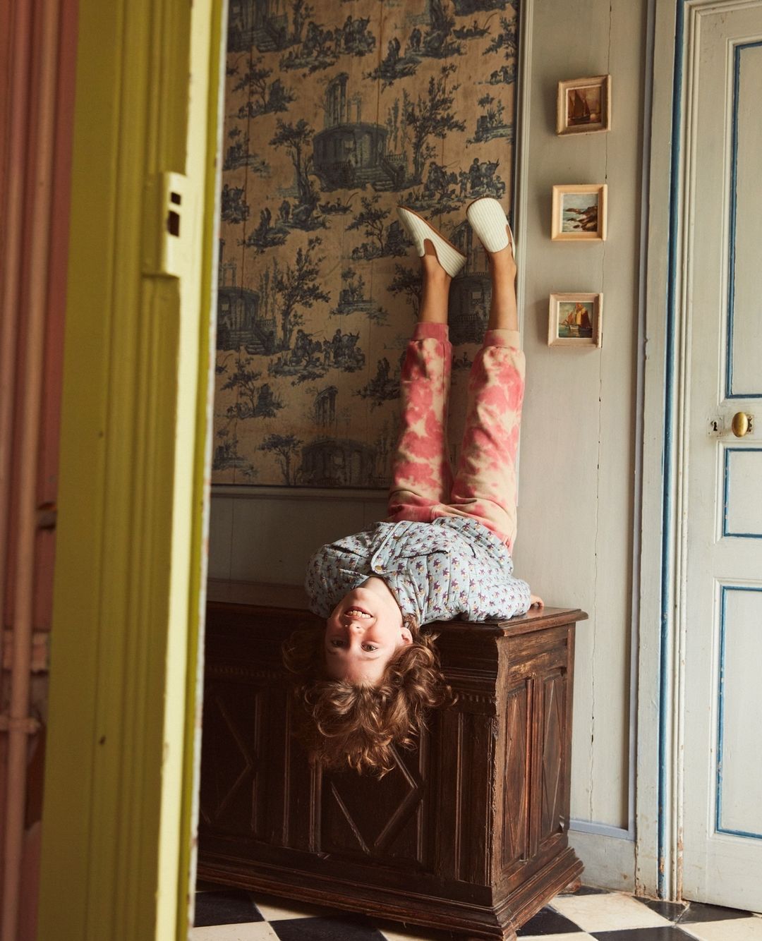 Upside-down view of a child balancing on a wooden chest, with patterned wallpaper, tiled floor, and an open doorway in background.