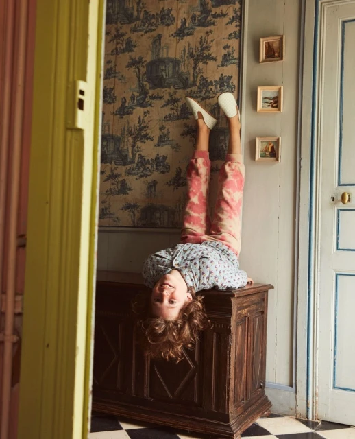 Upside-down view of a child balancing on a wooden chest, with patterned wallpaper, tiled floor, and an open doorway in background.