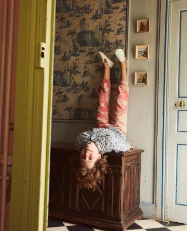 Upside-down view of a child balancing on a wooden chest, with patterned wallpaper, tiled floor, and an open doorway in background.