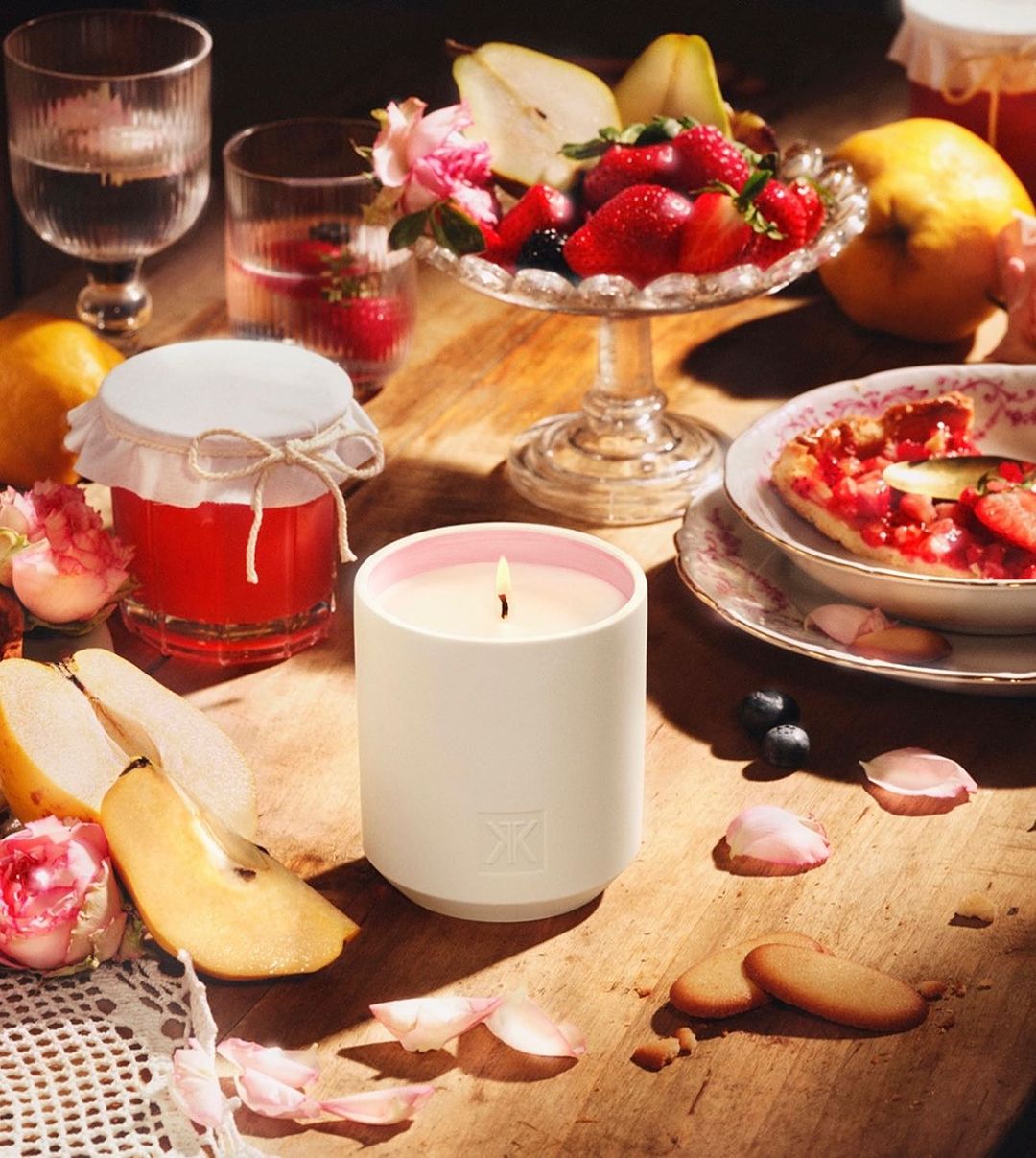 Still life with a lit candle surrounded by fresh fruit, jam, cookies, and flower petals on a wooden surface, creating a warm, inviting scene.