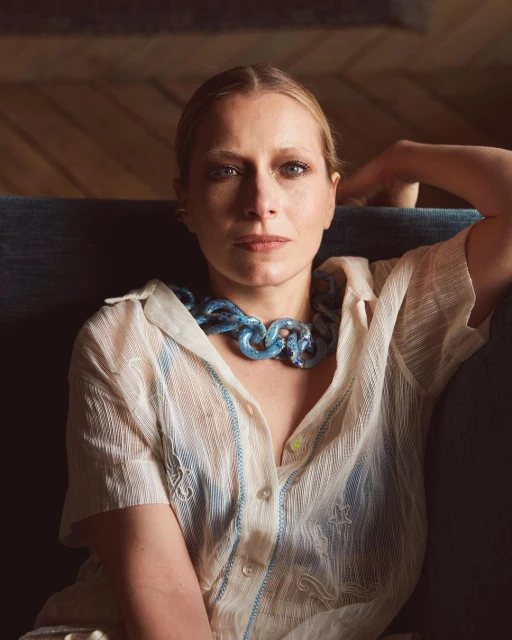 Portrait of a woman wearing a blue chunky necklace and sheer white top, seated against a blue couch with a soft, diffused lighting.