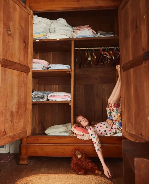 Red-haired child lying in a wooden armoire with open doors, wearing a polka dot top and floral shorts. Soft light fills the room.