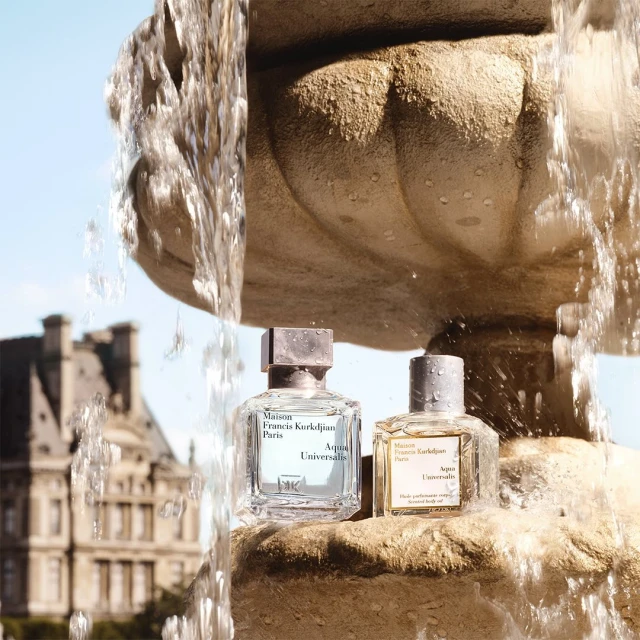 Two Maison Francis Kurkdjian Paris fragrance bottles sit on a stone fountain with flowing water, set against a backdrop of a building.