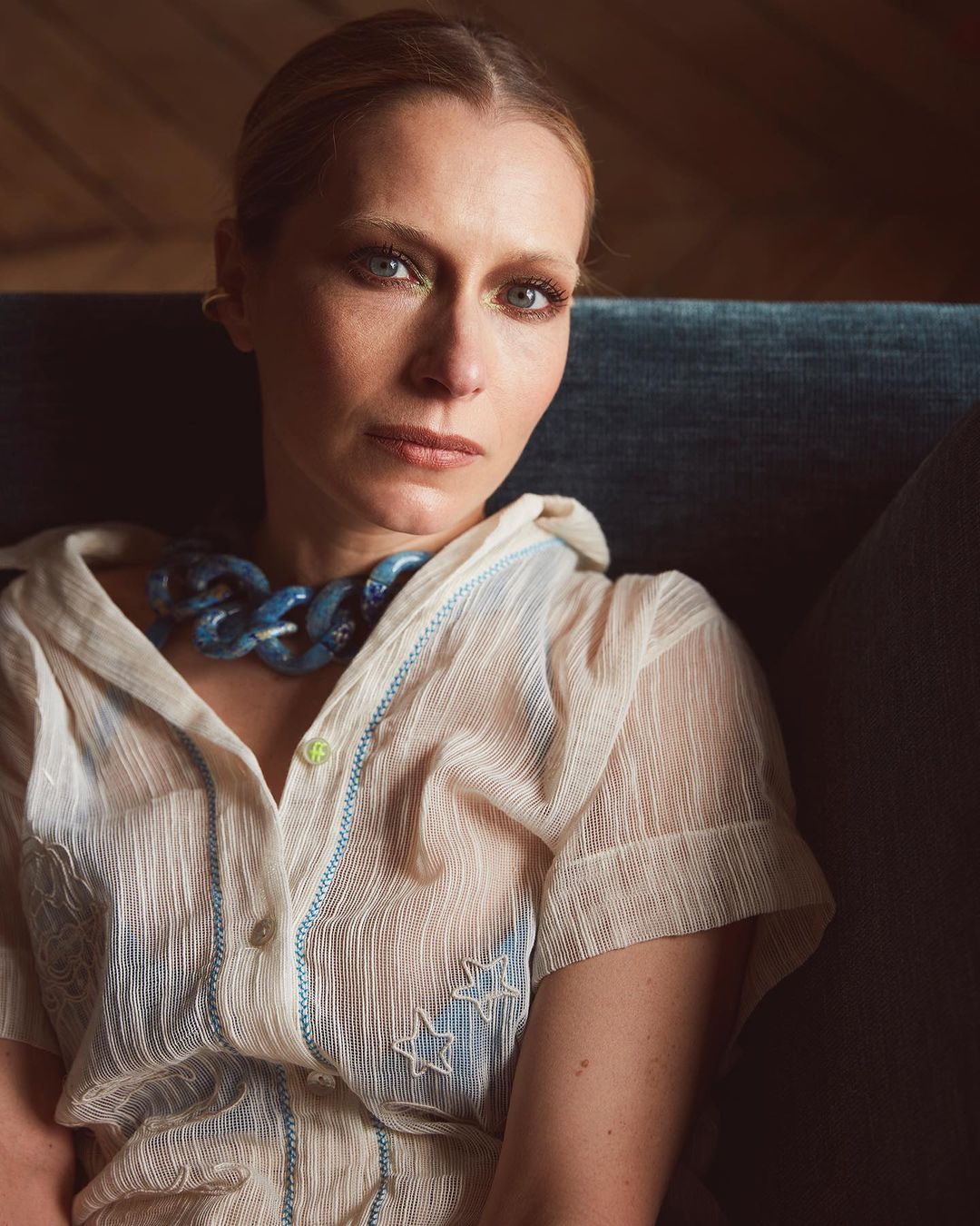 Portrait of a blond woman wearing a light blouse and a blue chunky necklace, sitting indoors near a window with natural light.
