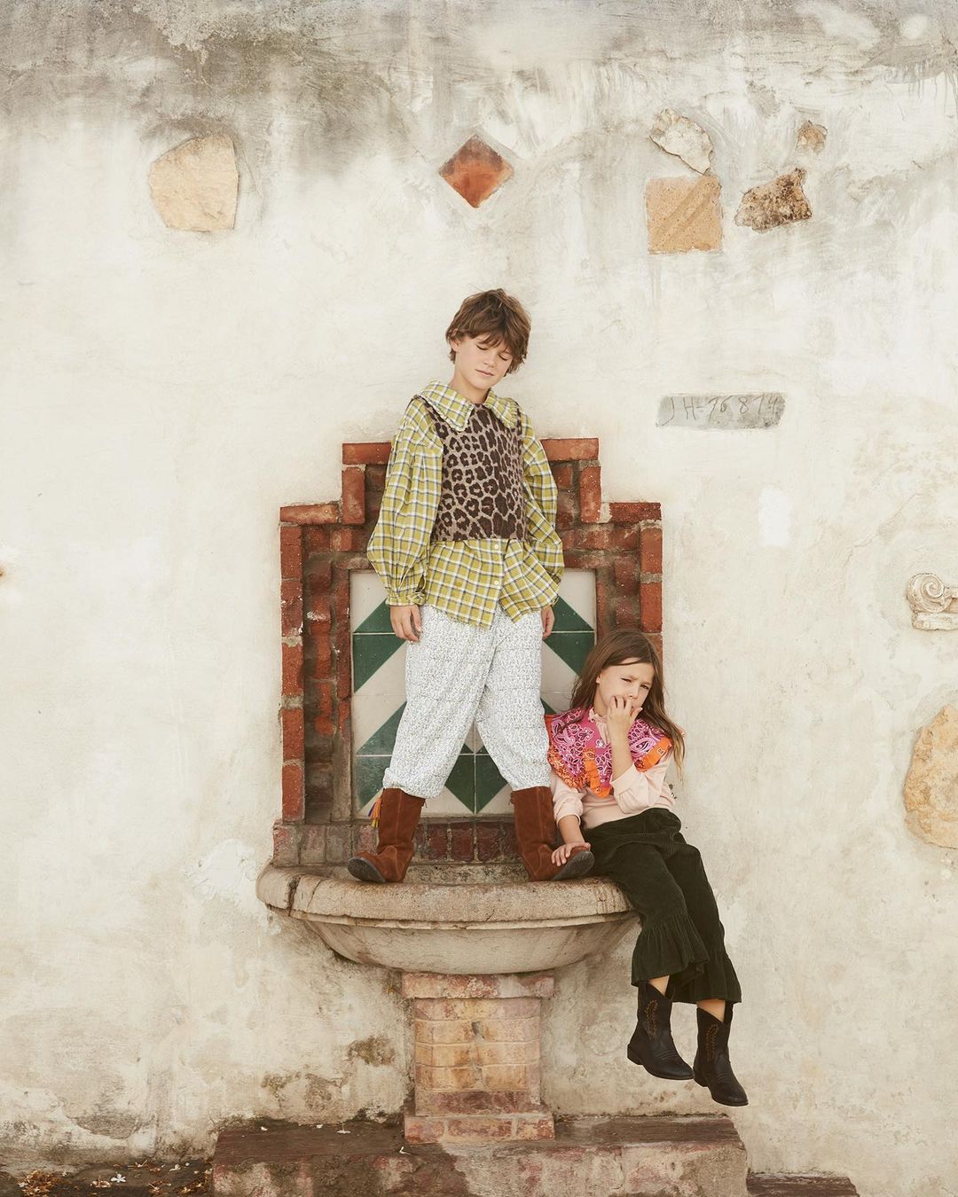 Two children pose outdoors on a weathered stone fountain in coordinated vintage-inspired clothing, against an aged stucco wall backdrop.