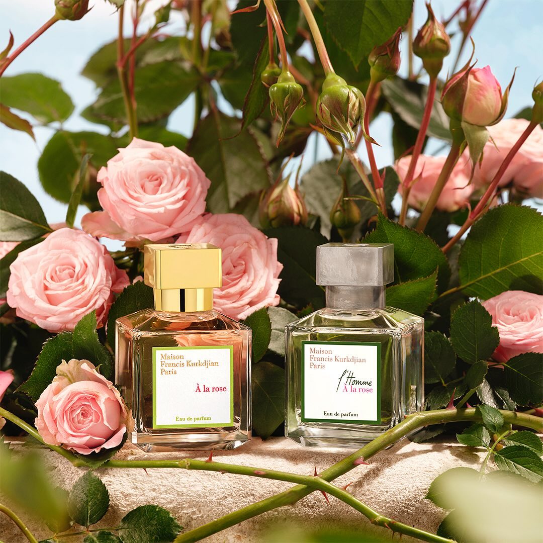 Two Maison Francis Kurkdjian À la rose perfumes are displayed amid light pink roses and greenery in an elegant, eye-level shot.
