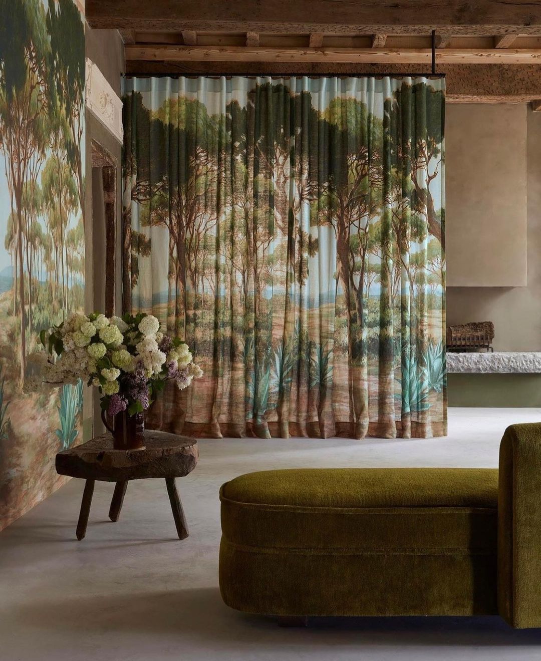 Olive green sofa and rustic wood table with white flowers in an interior space featuring curtain with landscape painting design.