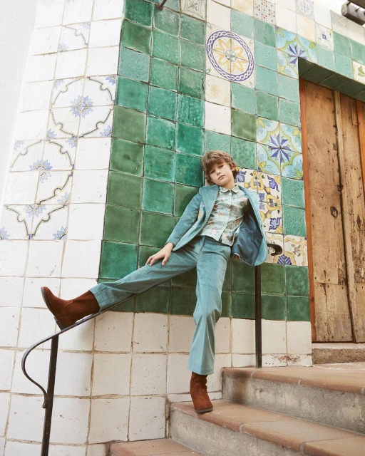 Stylish young adult poses on stairs in green suit, boots, and decorative-tile backdrop; fashion and architecture meet in this striking shot.