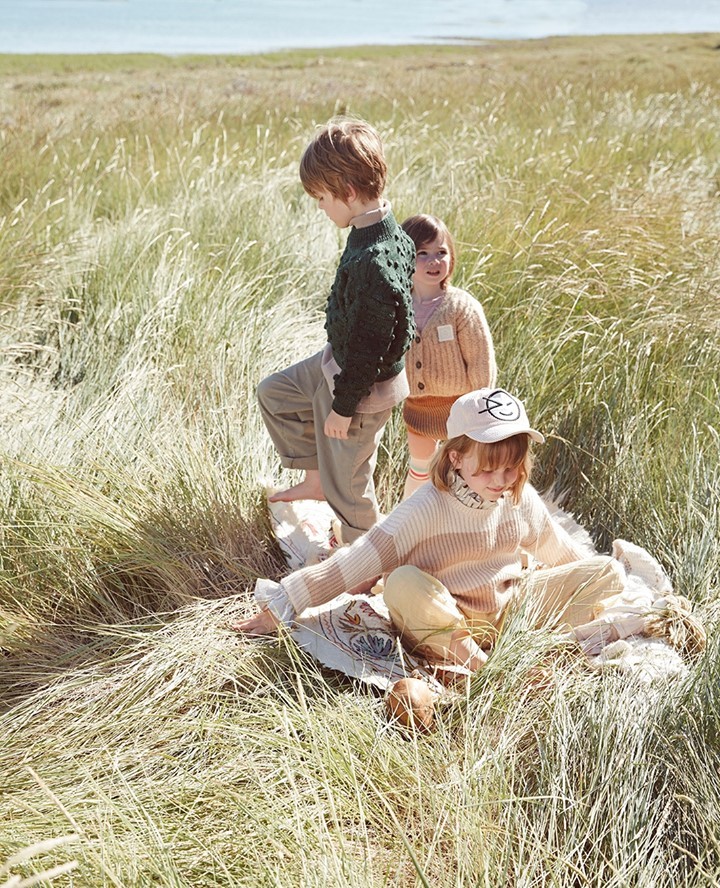 Three children pose outdoors in a grassy field, two standing and one sitting, wearing sweaters and a hat with a smiley face.
