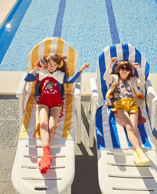Two children wearing colorful vintage-style outfits lounge on striped beach chairs next to a swimming pool on a sunny day.
