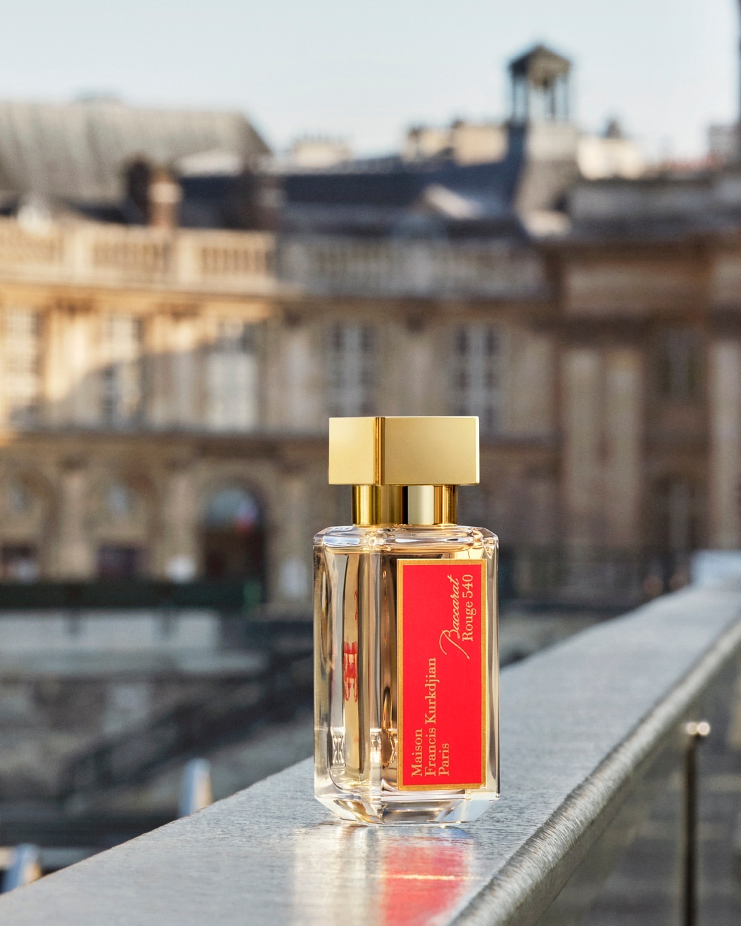 Baccarat Rouge 540 perfume bottle with golden cap on a metal railing with a blurry European city building in the background.