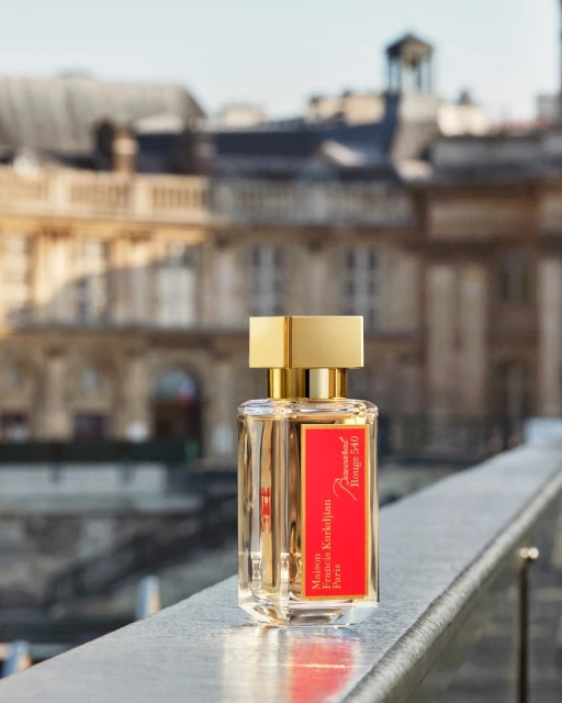 Baccarat Rouge 540 perfume bottle with golden cap on a metal railing with a blurry European city building in the background.