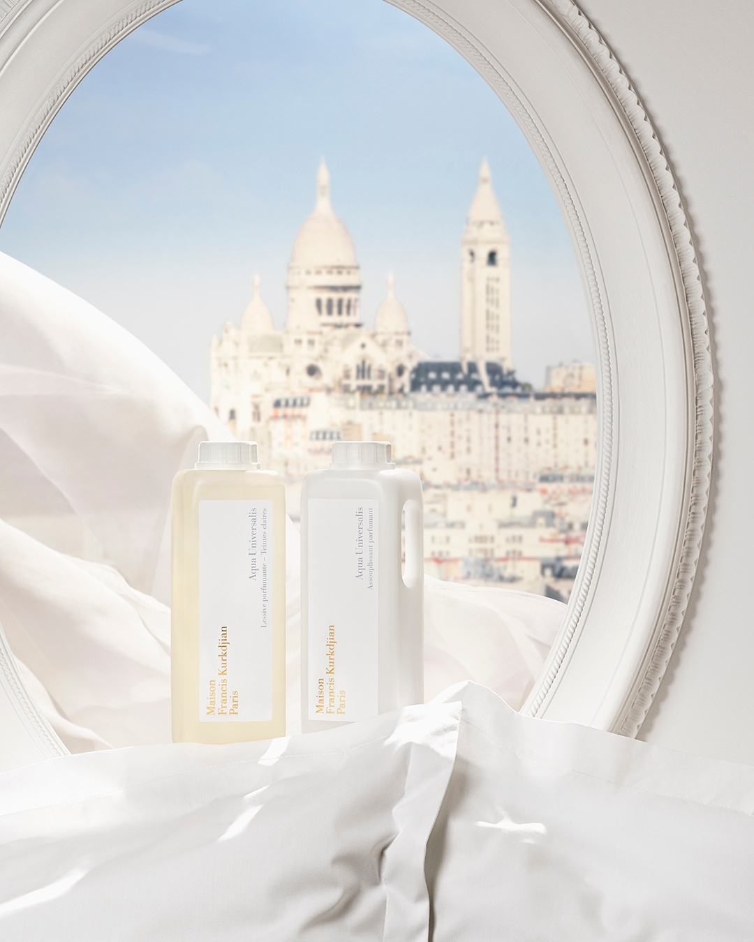 Two bottles of Maison Francis Kurkdjian detergent displayed on a white bed, with a view of Sacré-Cœur Basilica through a window.