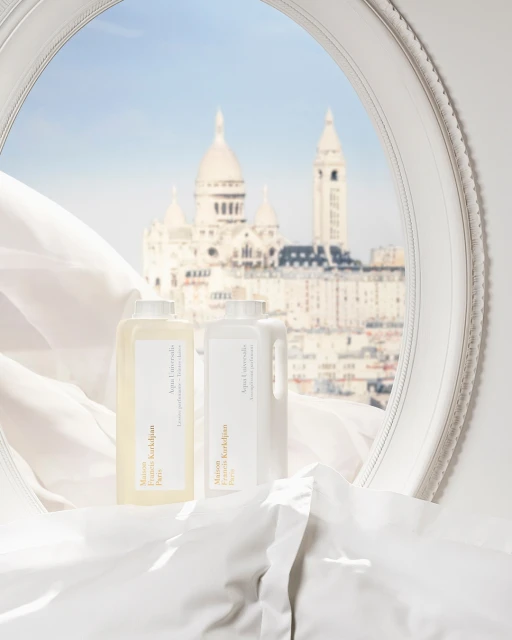 Two bottles of Maison Francis Kurkdjian detergent displayed on a white bed, with a view of Sacré-Cœur Basilica through a window.