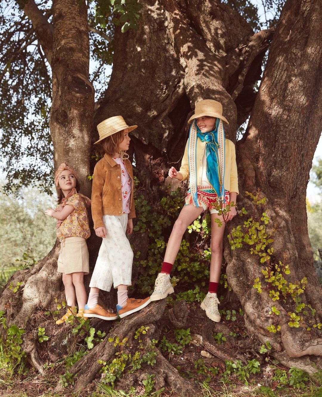 Three children pose outdoors near a large tree; two wear straw hats, colorful patterned outfits, and boots; one wears a headscarf and skirt.