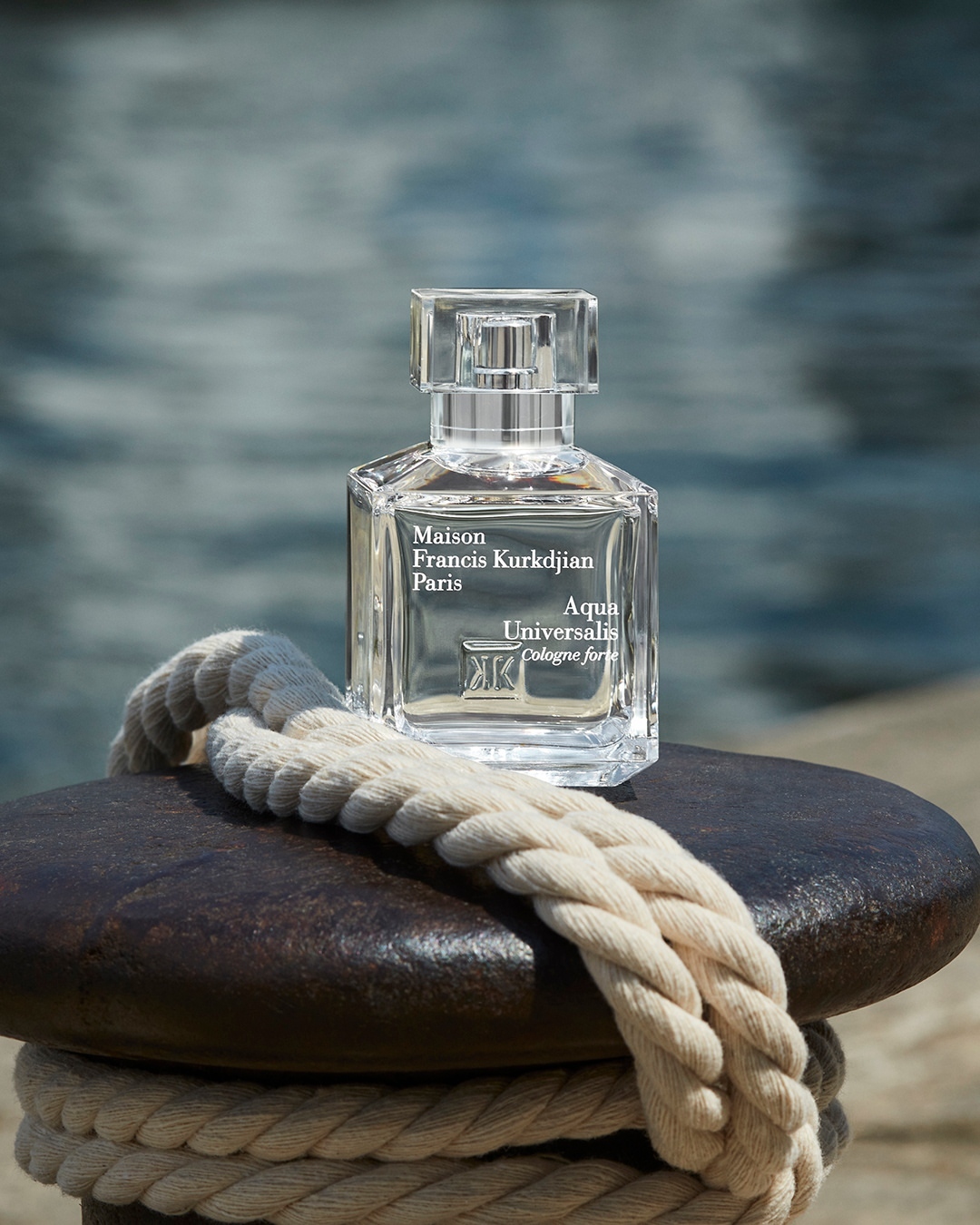 Crystal bottle of Maison Francis Kurkdjian Aqua Universalis cologne on a dark metal surface with a thick nautical rope. 