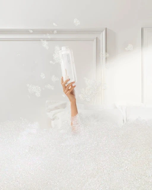 Arm holding white bottle in a white bubble bath against a white background with a white door frame for bath and beauty advertising.
