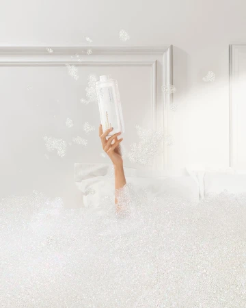 Arm holding white bottle in a white bubble bath against a white background with a white door frame for bath and beauty advertising.