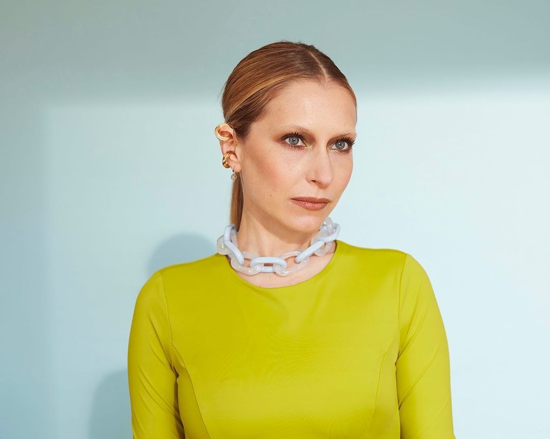 Portrait of a blonde adult woman in a chartreuse top and statement necklace against a light blue background.