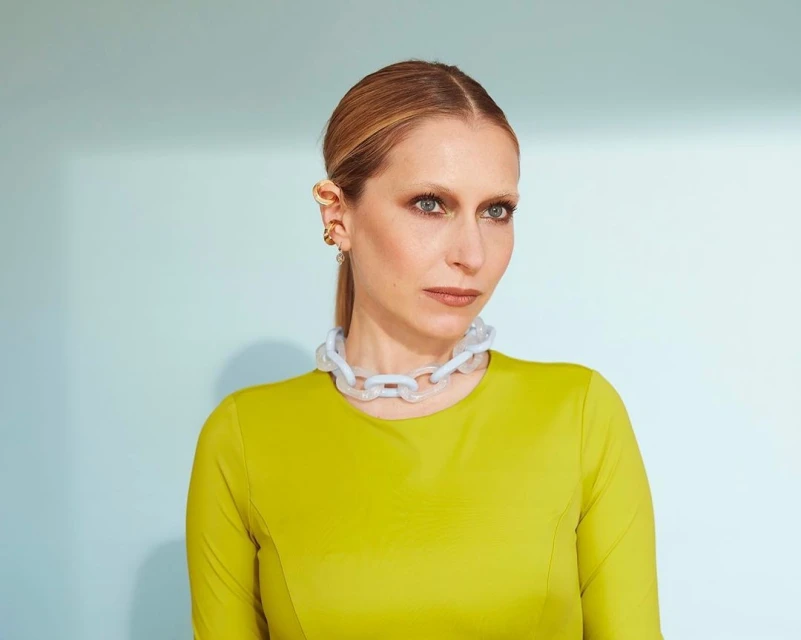 Portrait of a blonde adult woman in a chartreuse top and statement necklace against a light blue background.