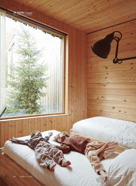 Two children lie on a twin bed in a wood-paneled bedroom with a view of a wintery tree. Lit by natural light and a black industrial lamp.