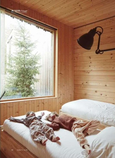 Two children lie on a twin bed in a wood-paneled bedroom with a view of a wintery tree. Lit by natural light and a black industrial lamp.