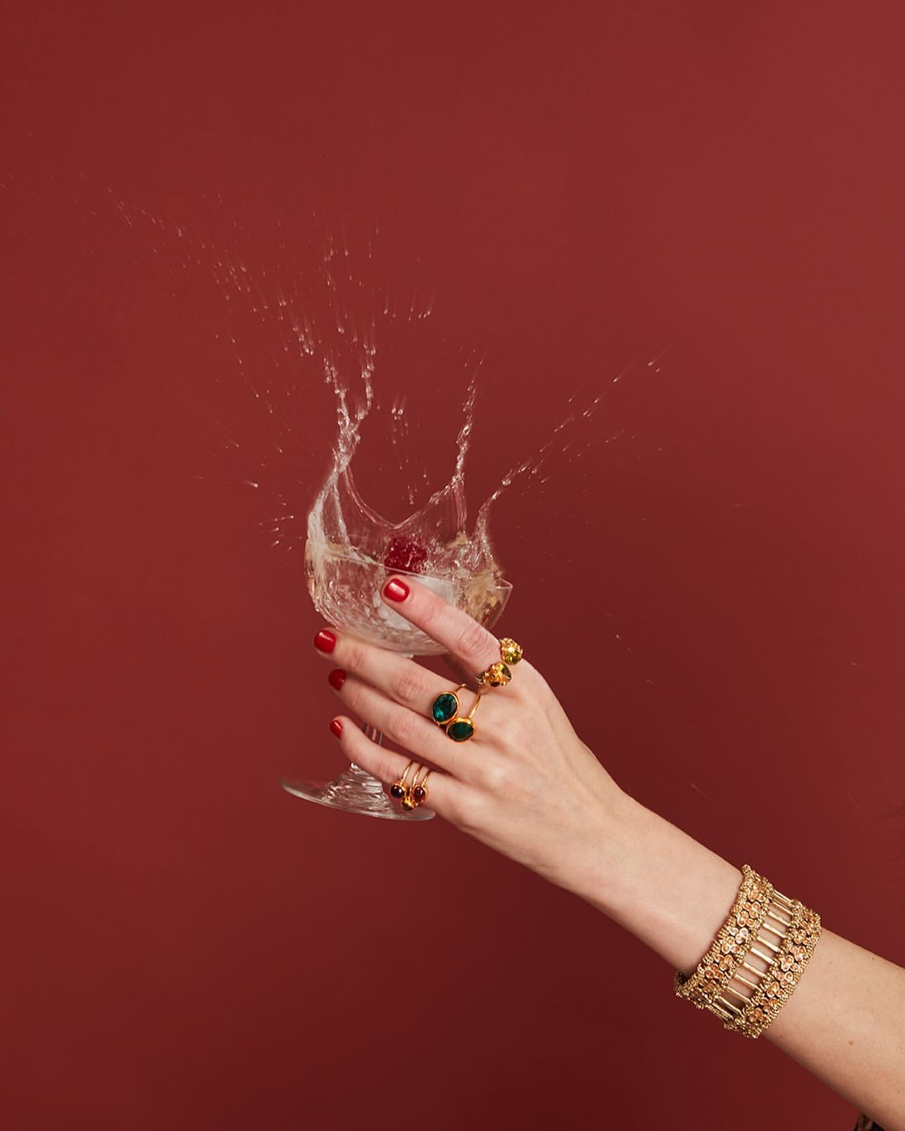 Elegant hand with red nails and gold jewelry holding a cocktail glass with splashing liquid against a solid brick-red backdrop.