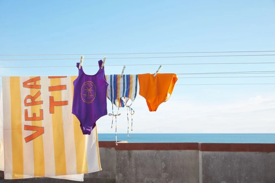 Outdoor shot of purple, orange, and striped swimsuits drying on a clothesline, with a striped towel, against a blue sky and ocean backdrop.