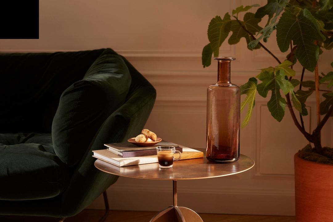 An espresso sits on a round table with a vase, books, and pastries, beside a green velvet couch and fig plant in a warm-toned interior.