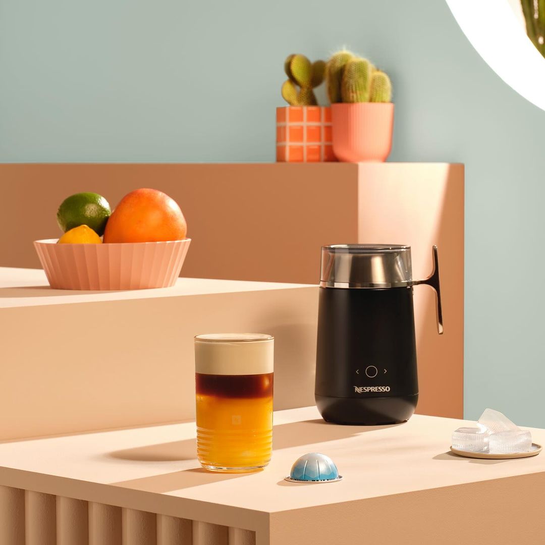 Still life shot of a layered drink, coffee machine, and citrus fruit on a multi-tiered peach platform against a light turquoise background.
