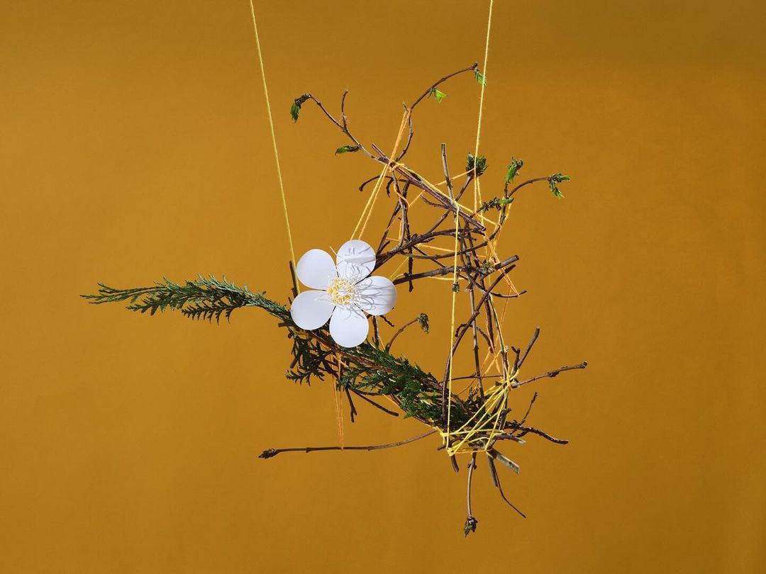 Botanical art installation features a white paper flower, green foliage, and bare branches suspended with yellow string against mustard background.