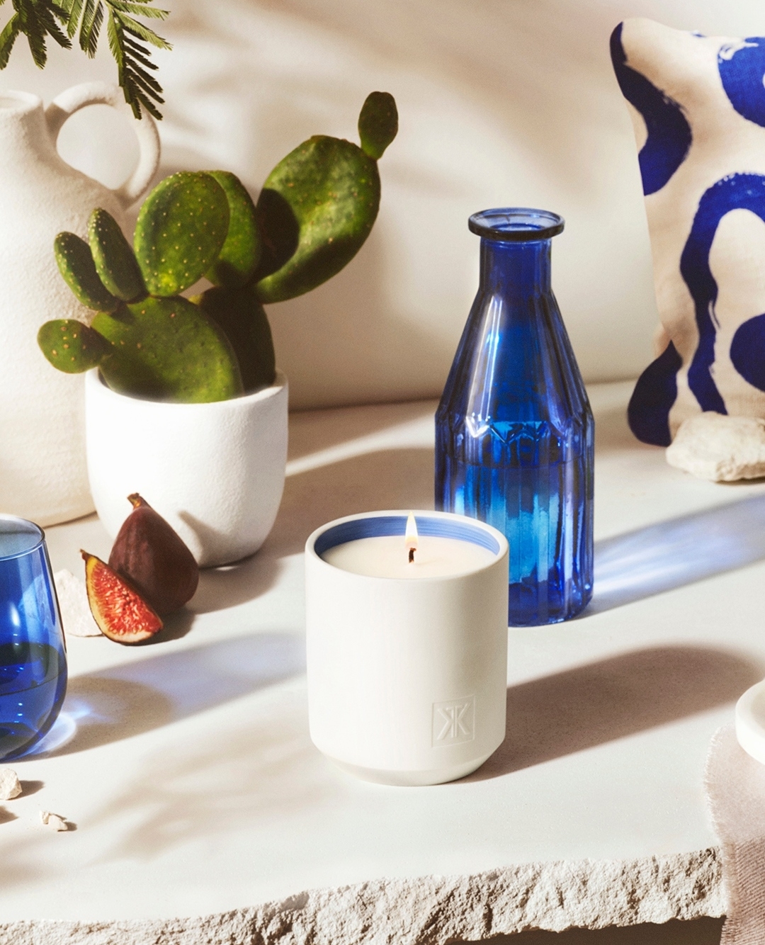 Still life captures a lit candle in a white cylindrical container, accompanied by a blue bottle, fresh figs, and a potted cactus.
