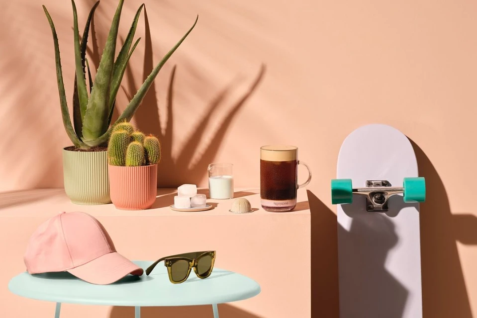 Still life featuring plants, drink, and skateboard against a peach backdrop with shadow patterns, evoking a summery, minimalist scene.