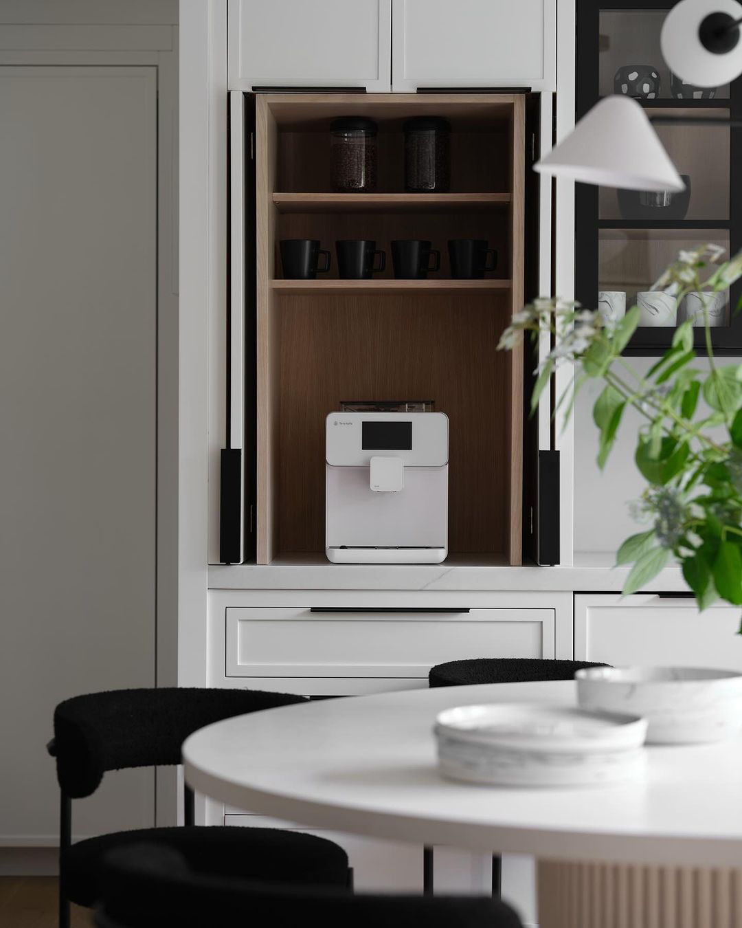 Modern kitchen interior with a white coffee machine inside a wooden cabinet, a white table, black chairs, and plants.