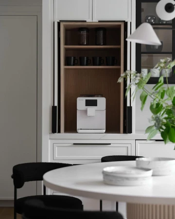 Modern kitchen interior with a white coffee machine inside a wooden cabinet, a white table, black chairs, and plants.