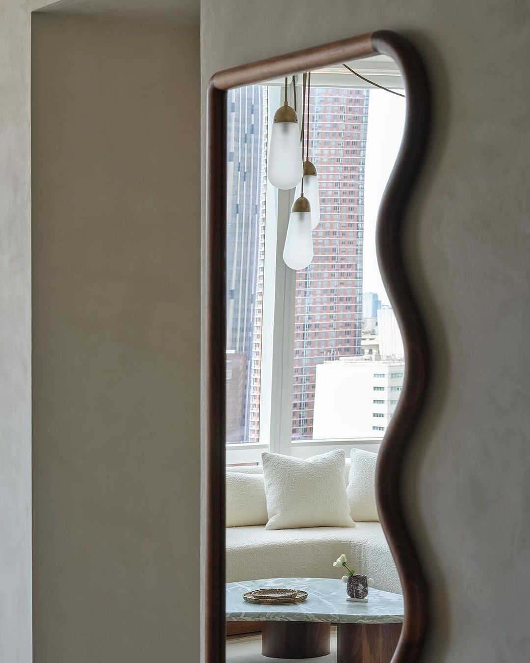 Interior view reflected in a modern mirror featuring a neutral palette, textured couch, marble table, and city view through the window.