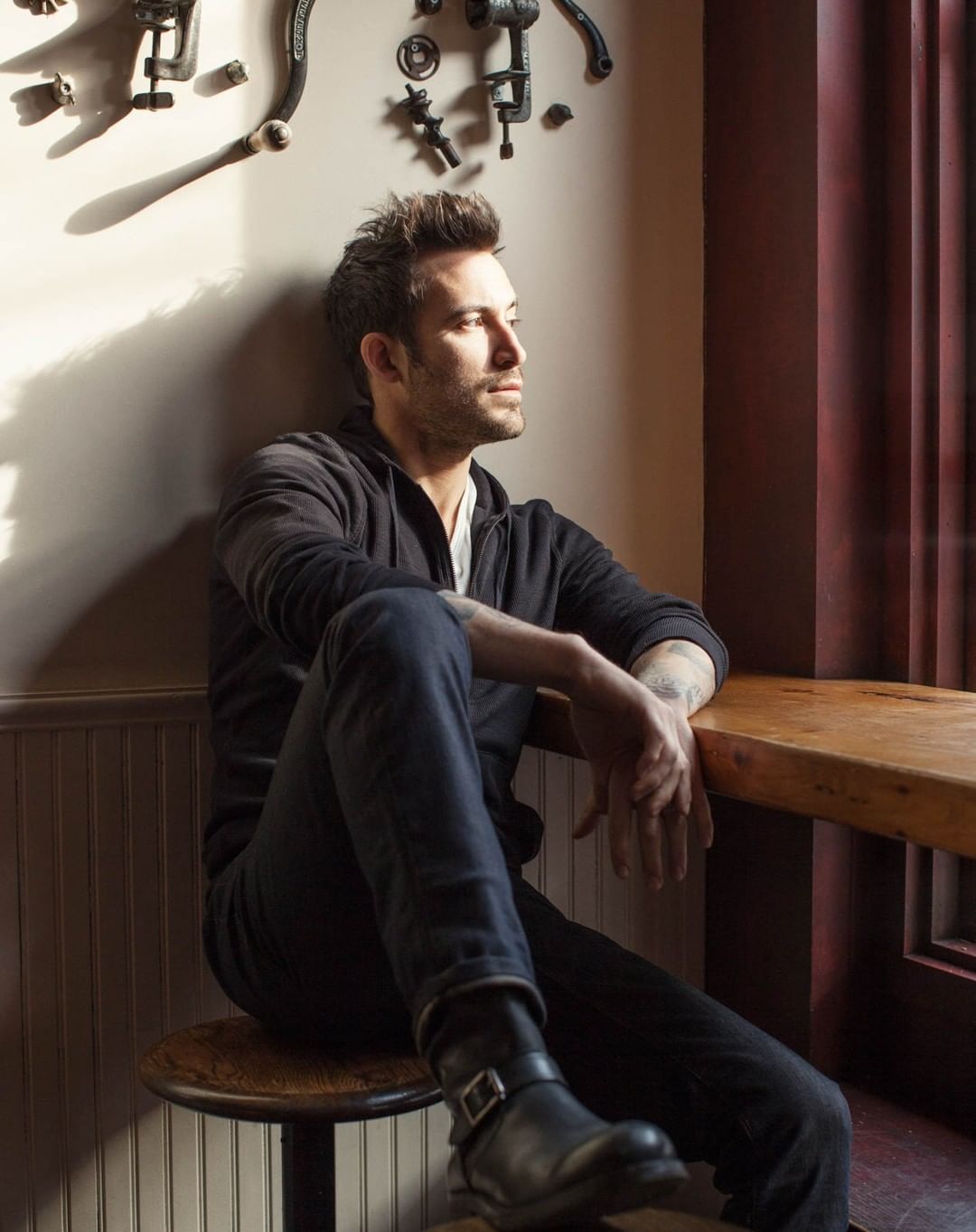 Adult male with beard and tattoos sits in an indoor cafe setting near a window and rustic decorations, wearing a dark hoodie and boots.