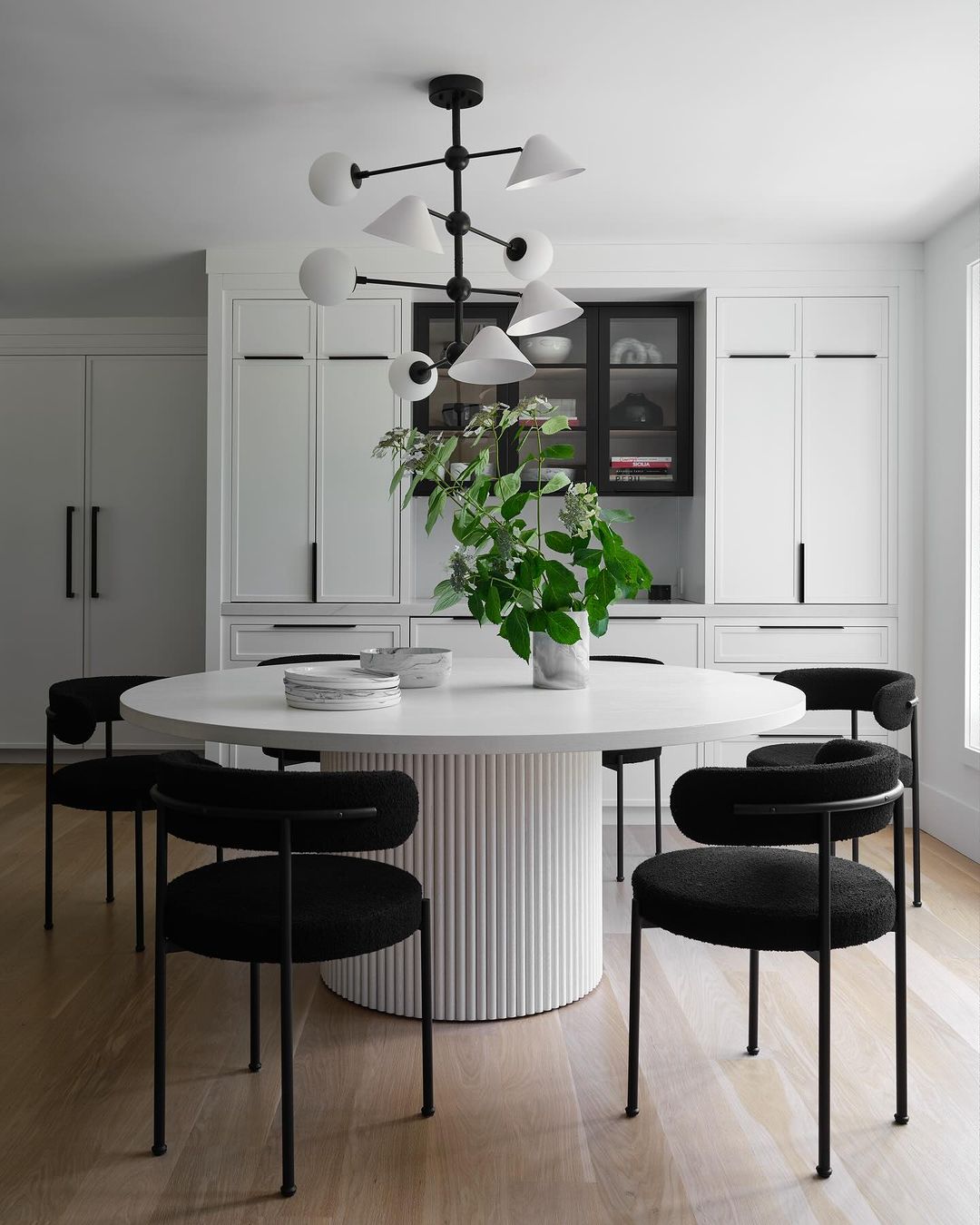 Modern dining room features a white round table, black upholstered chairs, a statement chandelier, and built-in cabinetry, creating a stylish space.